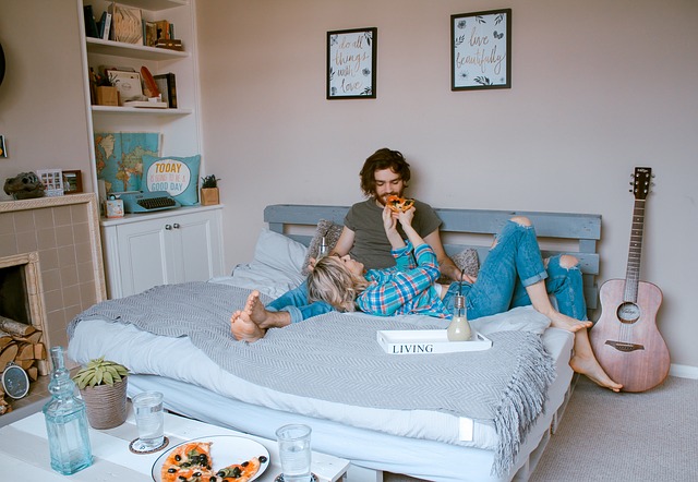 room couple in bed together eating pizza, girl is feeding guy pizza, lying on his lap